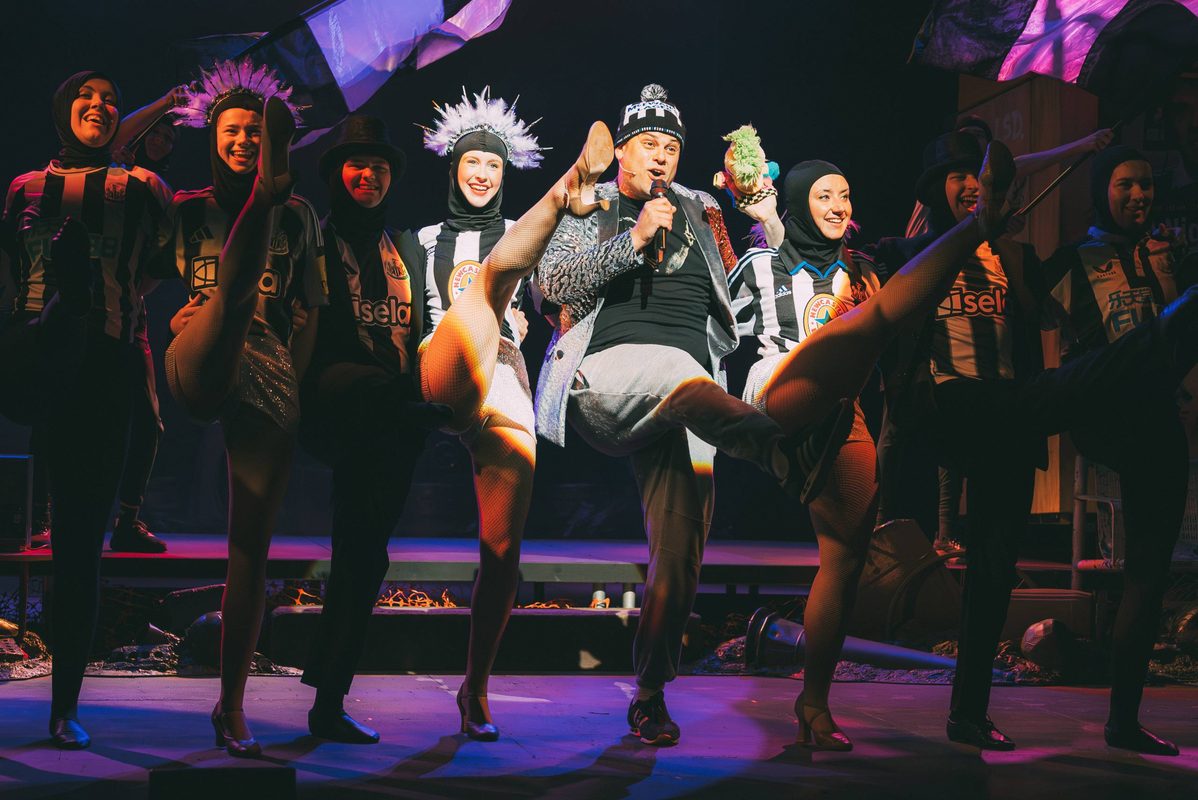 The company high-kicking in Newcastle shirts and sequins while a performer sings into a microphone