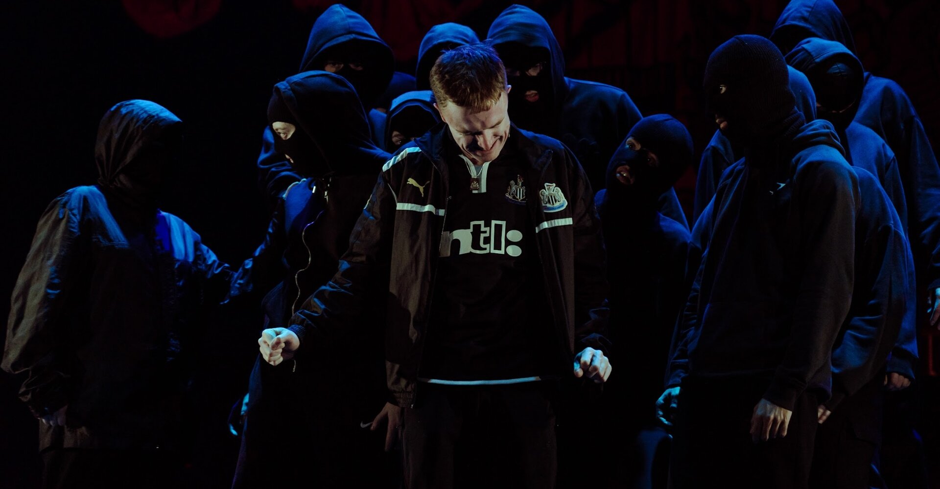 Gerry in a Newcastle United shirt surrounded by hooded figures under blue stage light